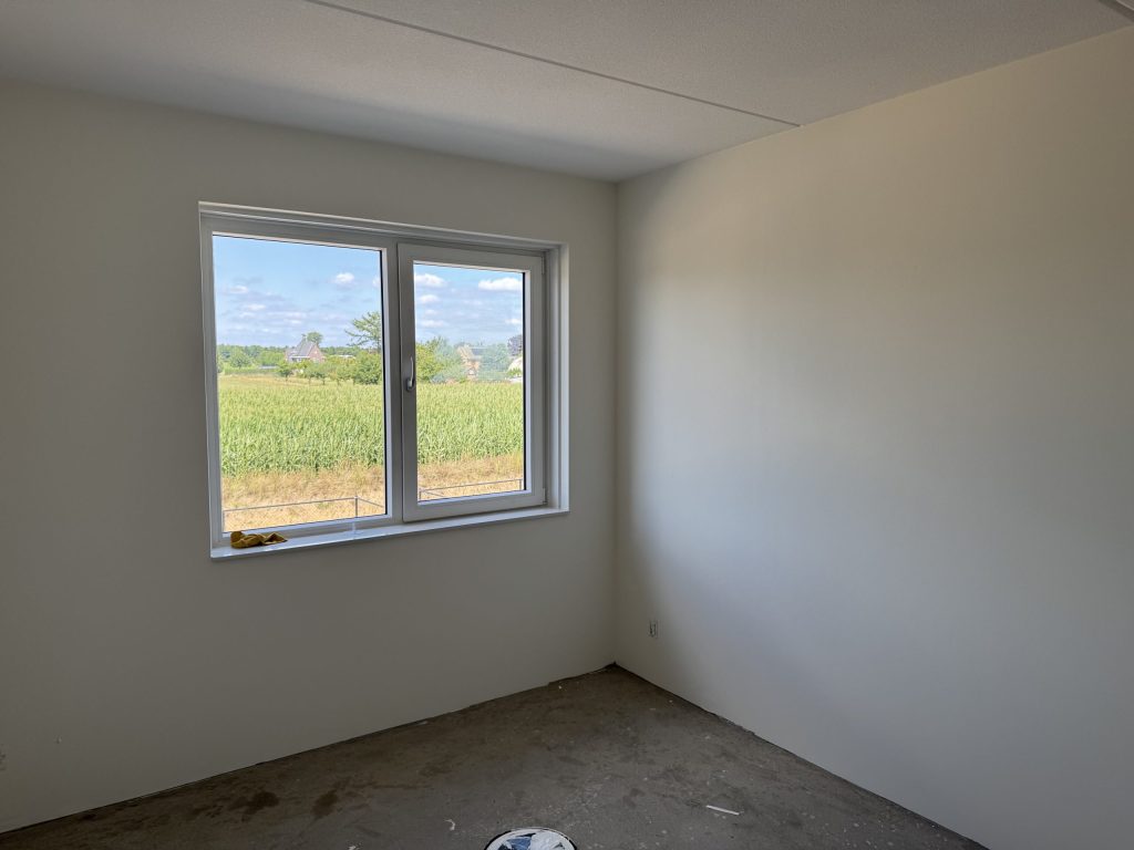 Lege slaapkamer met net gesausde witte vlieswanden en een raam dat uitkijkt op een maisveld