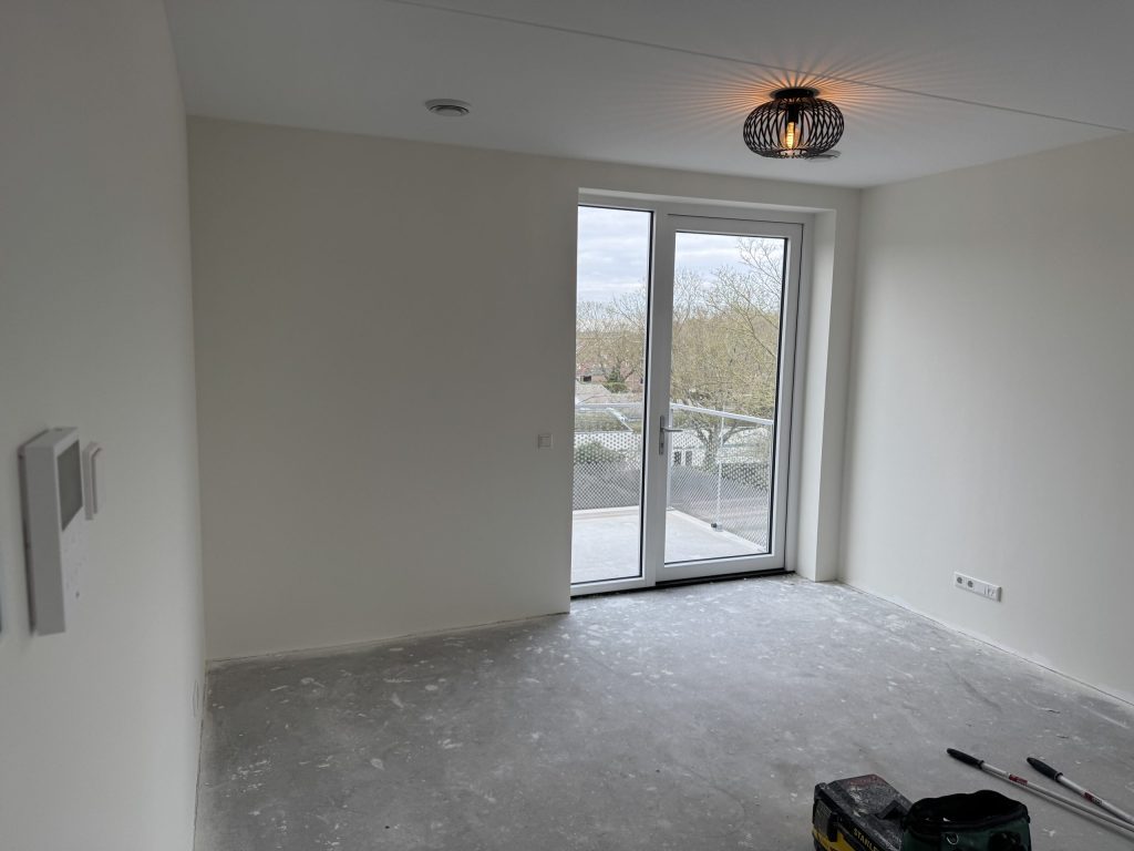Lichte kamer met balkondeuren en witte vlieswanden, rollers en gereedschapstas nog op de betonvloer