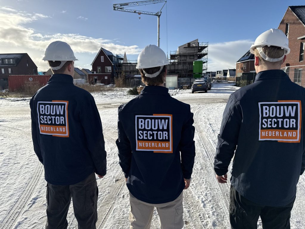 Drie vakmensen van Bouwsector Nederland met witte helm op een besneeuwde bouwstraat met huizen in aanbouw